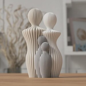 Puede incluir: Un conjunto de esculturas familiares, con dos figuras grandes y dos peque&ntilde;as. Las figuras est&aacute;n talladas con l&iacute;neas verticales, creando un efecto rayado. Las dos figuras m&aacute;s grandes son de color blanquecino, mientras que las m&aacute;s peque&ntilde;as son grises. La escultura se exhibe sobre una superficie de madera.