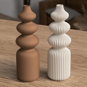 May include: Two decorative vases. One is a light brown color with a textured surface, and the other is white with vertical ribbed detailing. Both have a stacked, bulbous design and are displayed on a wooden surface.