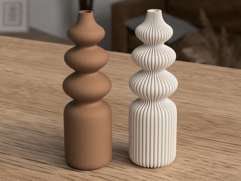 May include: Two decorative vases. One is a light brown color with a textured surface, and the other is white with vertical ribbed detailing. Both have a stacked, bulbous design and are displayed on a wooden surface.