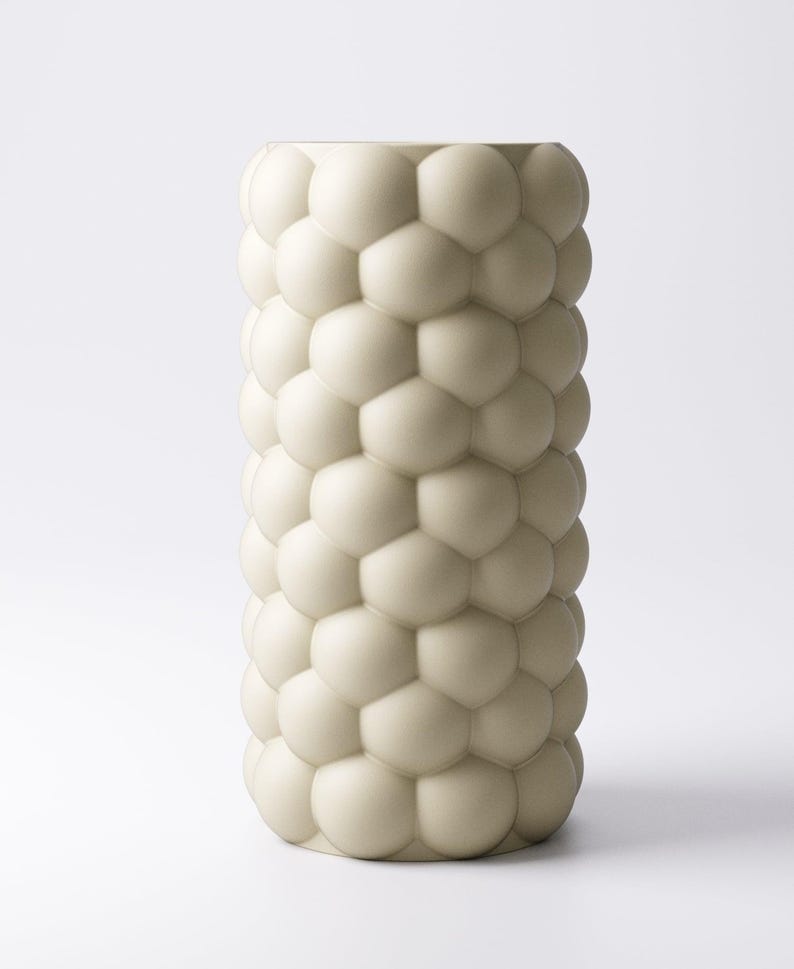 May include: A tall, off-white vase with a textured surface. The vase is cylindrical and covered in a pattern of rounded, interconnected shapes, resembling bubbles or cells. The design gives the vase a unique, modern aesthetic.