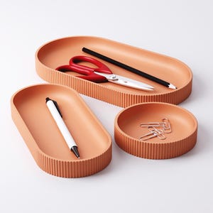 May include: A set of three terracotta-colored desk organizers. The set includes an oval tray with red scissors and a black pencil, a smaller oval tray with a white pen, and a round tray with paper clips.