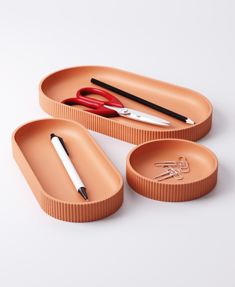 May include: A set of three terracotta-colored desk organizers. The set includes an oval tray with red scissors and a black pencil, a smaller oval tray with a white pen, and a round tray with paper clips.