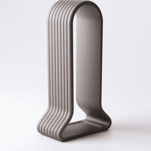 May include: A modern, minimalist sculpture in a muted gray color. The design features a curved, U-shaped form with vertical, ridged lines on one side, creating a textured effect. The sculpture stands on a flat base.