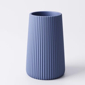 May include: A blue vase with a ribbed exterior. The vase has a cylindrical shape that tapers slightly towards the base. The color is a muted blue, and the surface has vertical ridges. The vase is likely made of ceramic or a similar material.