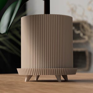 Puede incluir: Una maceta cil&iacute;ndrica de color marr&oacute;n claro con ranuras verticales. La maceta se apoya sobre una base a juego con tres peque&ntilde;os pies c&oacute;nicos blancos. Se ve una hoja verde de planta, lo que sugiere su uso para plantas de interior.