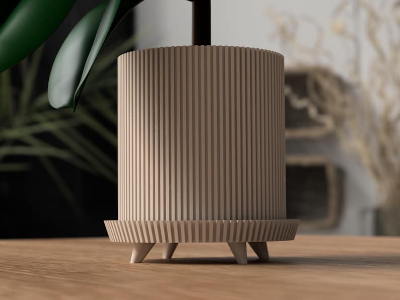 Puede incluir: Una maceta cil&iacute;ndrica de color marr&oacute;n claro con ranuras verticales. La maceta se apoya sobre una base a juego con tres peque&ntilde;os pies c&oacute;nicos blancos. Se ve una hoja verde de planta, lo que sugiere su uso para plantas de interior.