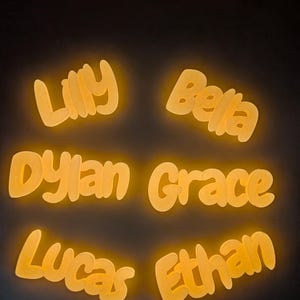 May include: Illuminated name signs in a warm yellow colour. The names "Lilly", "Bella", "Dylan", "Grace", "Lucas", and "Ethan" are presented in a playful, rounded font, ideal for nursery or bedroom decoration.