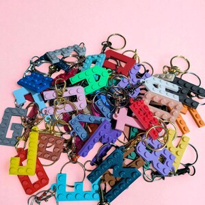 May include: A collection of colorful letter keychains in various colors, including blue, green, red, and purple. Each letter is designed to resemble interlocking toy bricks and is attached to a metal keyring. The keychains are arranged on a pink background.