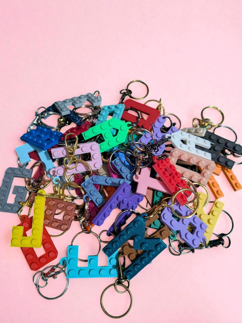 May include: A collection of colorful letter keychains in various colors, including blue, green, red, and purple. Each letter is designed to resemble interlocking toy bricks and is attached to a metal keyring. The keychains are arranged on a pink background.