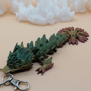 May include: A detailed, multi-colored dragon keychain. The dragon is primarily green and brown, with a metallic silver keychain ring and clasp. The dragon's body is segmented, with a textured, spiky appearance. The background features white crystal formations.