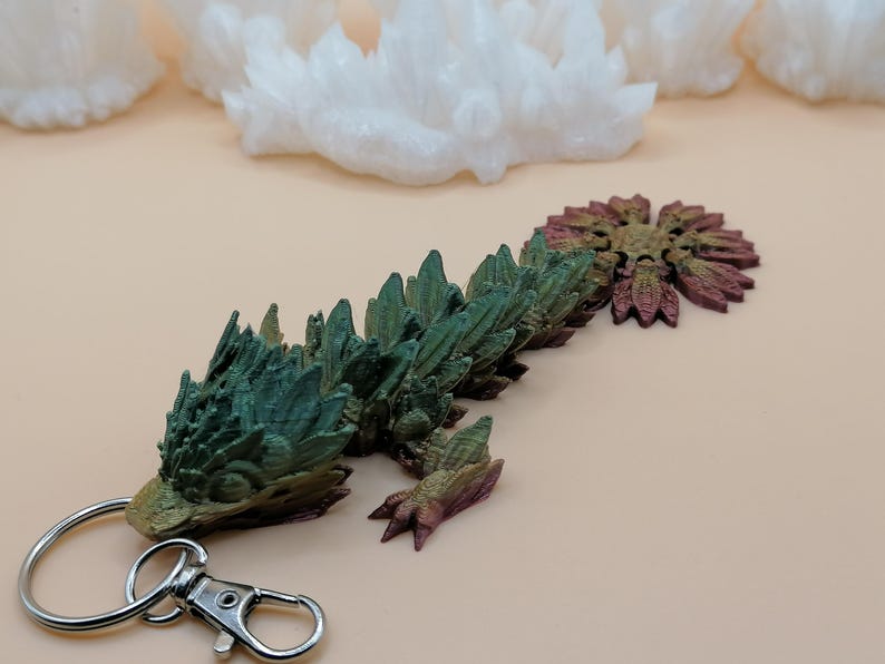 May include: A detailed, multi-colored dragon keychain. The dragon is primarily green and brown, with a metallic silver keychain ring and clasp. The dragon's body is segmented, with a textured, spiky appearance. The background features white crystal formations.