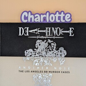 May include: A black book titled Death Note with silver lettering and a silver skull graphic, partially covered by a light beige book titled Another Note The Los Angeles BB Murder Cases with silver text, and a purple name tag reading Charlotte.