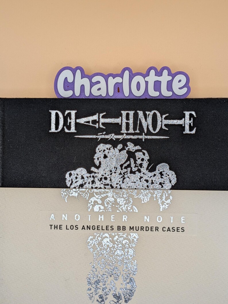 May include: A black book titled Death Note with silver lettering and a silver skull graphic, partially covered by a light beige book titled Another Note The Los Angeles BB Murder Cases with silver text, and a purple name tag reading Charlotte.