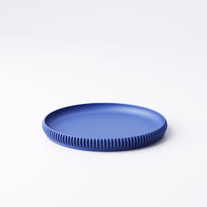 May include: A round, cobalt blue plate with a ribbed edge. The plate is made of a solid material and sits on a white background. The plate's design is simple and modern, suitable for serving food.