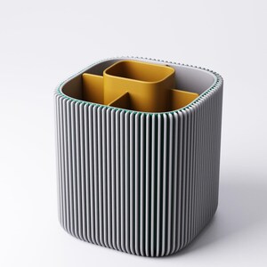 May include: A contemporary desk tidy with a grey, ribbed outer shell and a mustard yellow interior. It features multiple compartments for pens, pencils, and other stationery items. The design is minimalist and functional, suitable for home or office use.