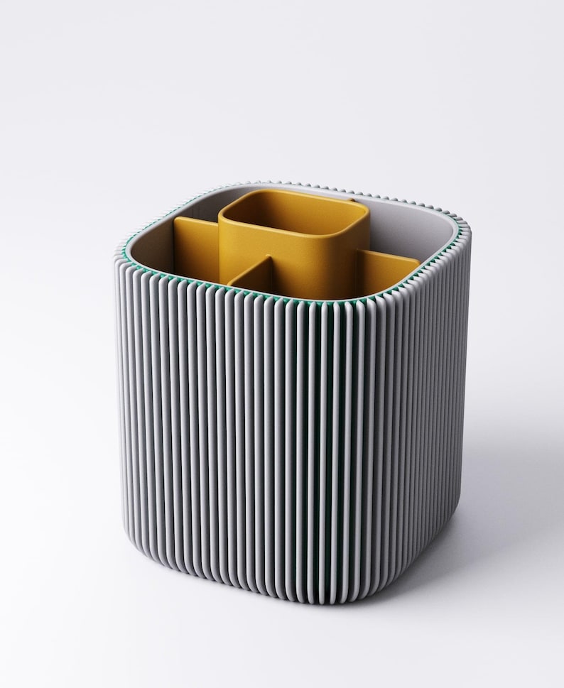 May include: A contemporary desk tidy with a grey, ribbed outer shell and a mustard yellow interior. It features multiple compartments for pens, pencils, and other stationery items. The design is minimalist and functional, suitable for home or office use.