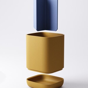 May include: A three-piece container set in a minimalist style. The set includes a mustard yellow, ribbed, square-shaped container with a matching lid and a blue, angular divider. The pieces are suspended in the air against a white background.