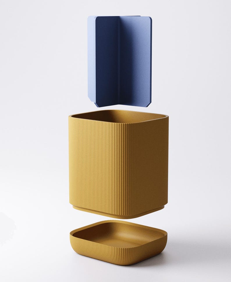 May include: A three-piece container set in a minimalist style. The set includes a mustard yellow, ribbed, square-shaped container with a matching lid and a blue, angular divider. The pieces are suspended in the air against a white background.