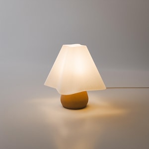 May include: A table lamp featuring a rounded, ribbed mustard-yellow base and a wide, pleated, off-white lampshade. The lamp emits a soft, warm glow from within, illuminating the surrounding area with a gentle light while resting on a flat surface.
