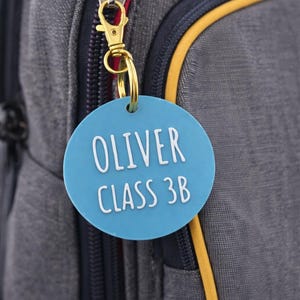 May include: A blue circular tag with the name "OLIVER" and "CLASS 3B" in white lettering, attached to a gold-colored keychain. The tag hangs from a gray backpack with a red zipper.
