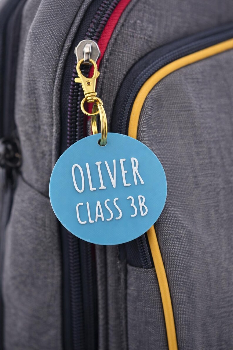 May include: A blue circular tag with the name "OLIVER" and "CLASS 3B" in white lettering, attached to a gold-colored keychain. The tag hangs from a gray backpack with a red zipper.