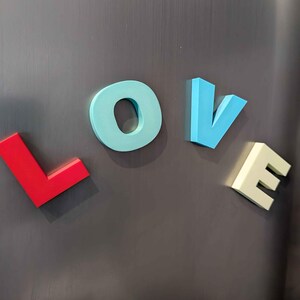 May include: Colorful magnetic letters spelling the word "LOVE" are displayed on a dark gray surface. The letters are in red, light blue, blue, and off-white, creating a vibrant and eye-catching display.