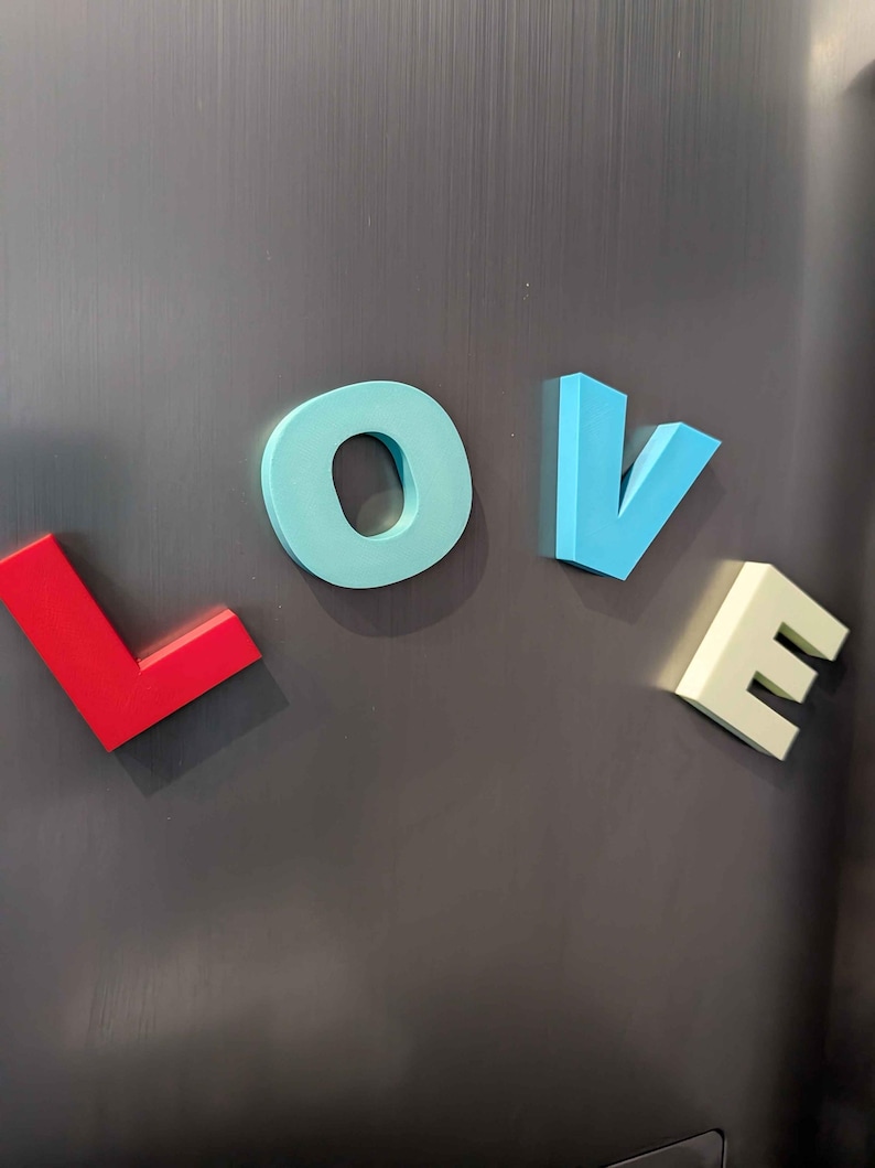 May include: Colorful magnetic letters spelling the word "LOVE" are displayed on a dark gray surface. The letters are in red, light blue, blue, and off-white, creating a vibrant and eye-catching display.