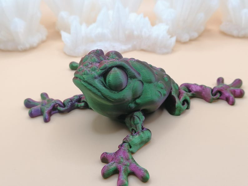 May include: A 3D-printed frog figurine in shades of green and purple. The frog has a textured skin and outstretched limbs. The figurine is set against a light background with white, crystal-like formations.