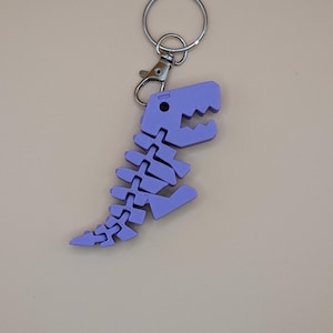 May include: A light purple dinosaur skeleton keychain. The keychain features a detailed, articulated design of a dinosaur skeleton, complete with a head, spine, and tail. It is attached to a silver keyring and a lobster clasp.