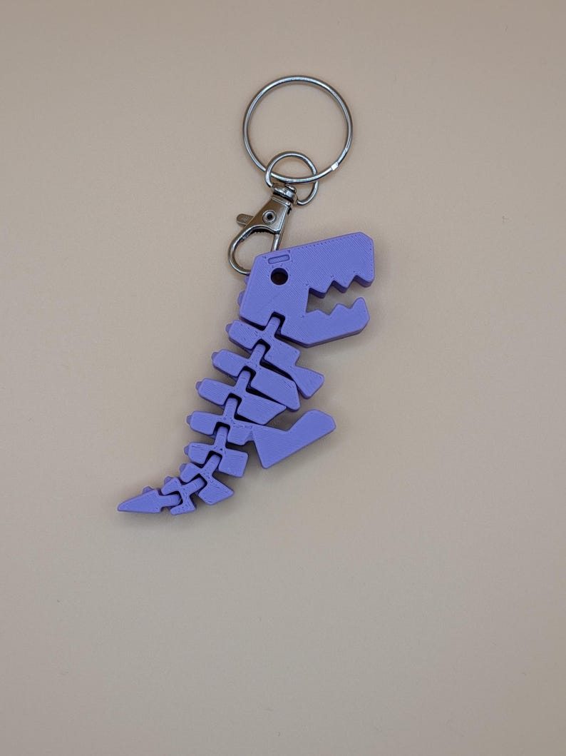 May include: A light purple dinosaur skeleton keychain. The keychain features a detailed, articulated design of a dinosaur skeleton, complete with a head, spine, and tail. It is attached to a silver keyring and a lobster clasp.