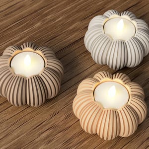 May include: Three decorative candle holders with a flower-like design. Each holder has a different color: beige, light gray, and peach. The holders each contain a lit, white, flameless candle, creating a warm ambiance.
