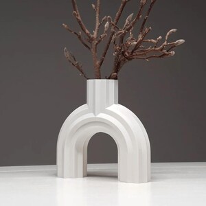 May include: A white, modern vase with an arched design. The vase has a rectangular neck and holds several brown branches. The vase is made of a solid material and sits on a white surface against a gray background.