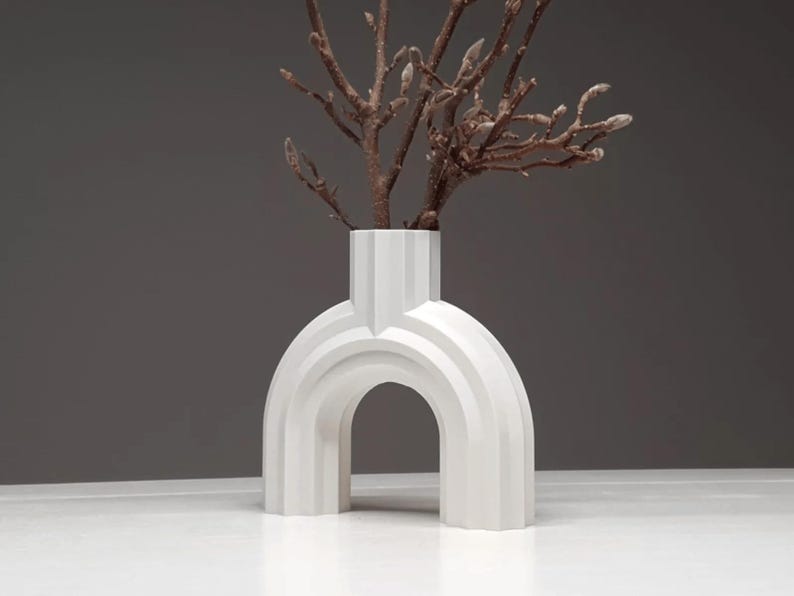 May include: A white, modern vase with an arched design. The vase has a rectangular neck and holds several brown branches. The vase is made of a solid material and sits on a white surface against a gray background.