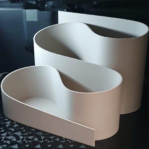 以下が含まれることがあります： さまざまなサイズの3つの白い、抽象的で湾曲した容器。 最大のものは背の高い円筒形で、中くらいのものは波のような形をしており、最小のものは浅い湾曲した形をしています。 オブジェクトは暗い表面にあります。
