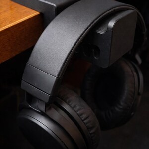 Puede incluir: Auriculares negros supraaurales colgando de un soporte de escritorio negro. Los auriculares tienen una diadema y almohadillas acolchadas. El soporte est&aacute; fijado a un escritorio de madera. La imagen es un primer plano.