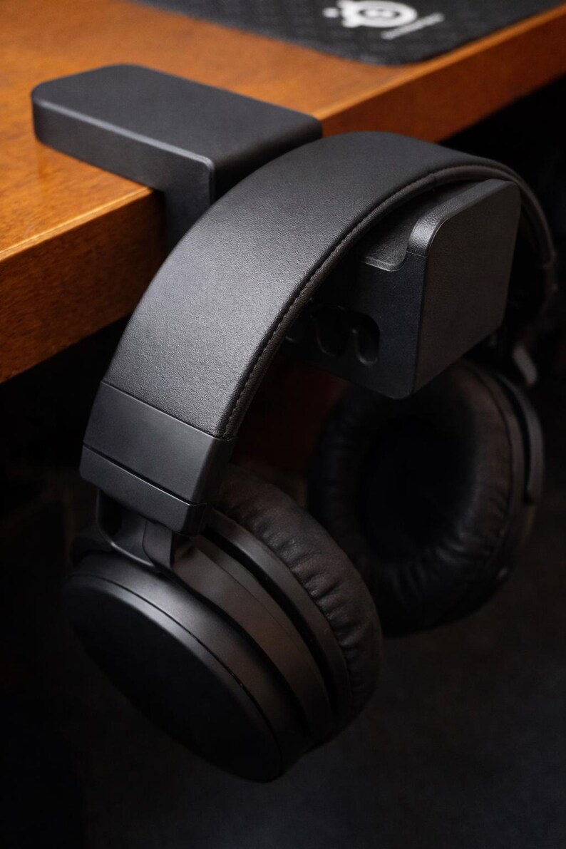 Puede incluir: Auriculares negros supraaurales colgando de un soporte de escritorio negro. Los auriculares tienen una diadema y almohadillas acolchadas. El soporte est&aacute; fijado a un escritorio de madera. La imagen es un primer plano.