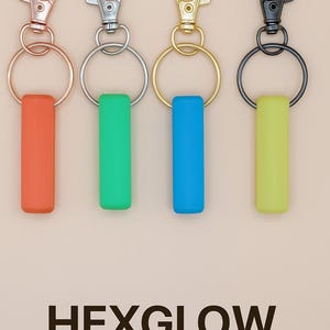 May include: Four keychains with colorful glow stick-shaped pendants. The keychains have different metal finishes: rose gold, silver, gold, and black. The pendants are orange, green, blue, and yellow. The text "HEXGLOW Glow Stick Keyring" is at the bottom.