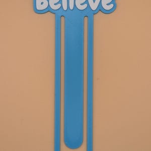 May include: A blue plastic bookmark shaped like a large paperclip with the word Believe written in white letters across the top. The bookmark has a textured surface and a long, slender vertical design that curves at the bottom end.