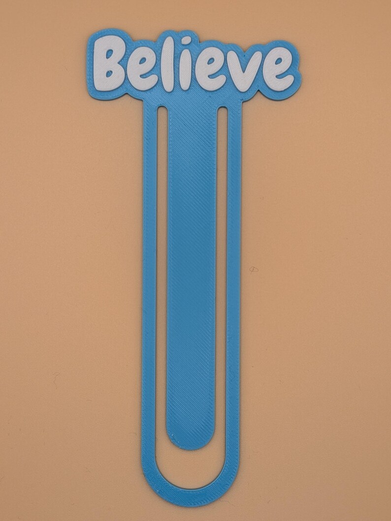 May include: A blue plastic bookmark shaped like a large paperclip with the word Believe written in white letters across the top. The bookmark has a textured surface and a long, slender vertical design that curves at the bottom end.