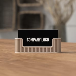 May include: A beige business card holder with vertical lines, holding a black business card with the words "COMPANY LOGO" in white. The holder sits on a wooden surface.