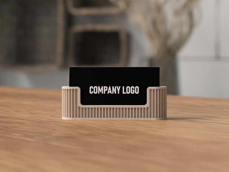 May include: A beige business card holder with vertical lines, holding a black business card with the words "COMPANY LOGO" in white. The holder sits on a wooden surface.