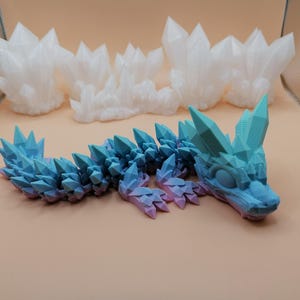 May include: A blue and purple articulated dragon toy with crystal-like spikes along its back and head. Three white crystal formations are in the background. The dragon is lying on a tan surface.