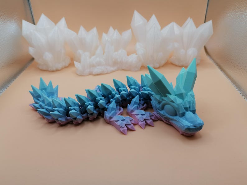 May include: A blue and purple articulated dragon toy with crystal-like spikes along its back and head. Three white crystal formations are in the background. The dragon is lying on a tan surface.