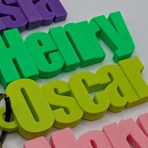May include: Close-up of colorful 3D-printed name keychains. The names "Henry" in green and "Oscar" in lime green are prominent. Other names, including "Isla" in purple and "Nora" in pink, are also visible. Each keychain has a metal ring.