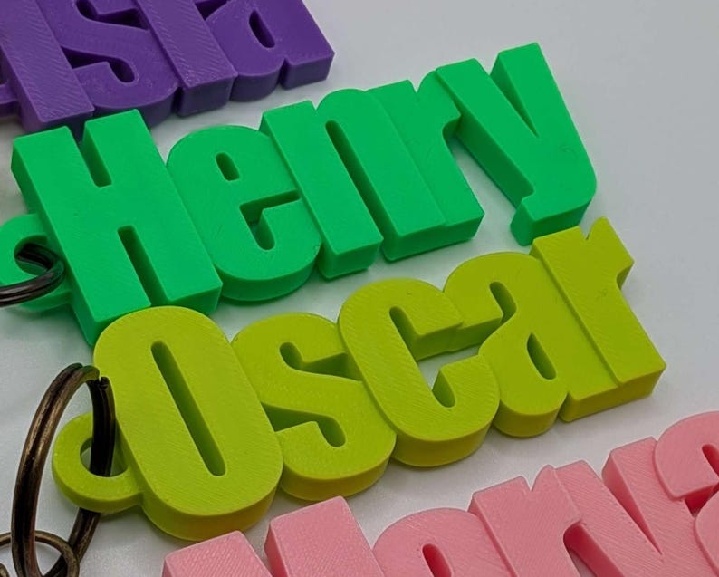 May include: Close-up of colorful 3D-printed name keychains. The names "Henry" in green and "Oscar" in lime green are prominent. Other names, including "Isla" in purple and "Nora" in pink, are also visible. Each keychain has a metal ring.