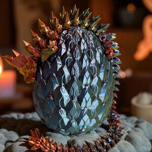 Op de afbeelding: Een decoratieve draken-eiersculptuur met metallic afwerking. Het ei is donkerblauw met een geometrisch patroon en heeft gouden en koperkleurige kristallen punten. Het staat op een getextureerd grijs oppervlak.