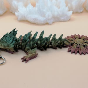 May include: A multi-colored, articulated dragon keychain. The dragon is primarily green and purple, with a silver key ring and clasp. The dragon's body is segmented, and the tail is a stylized flower shape.