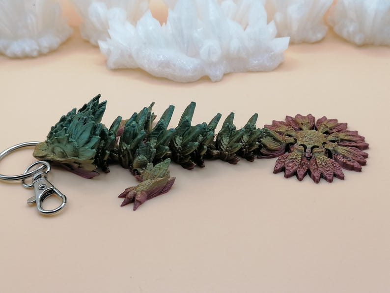 May include: A multi-colored, articulated dragon keychain. The dragon is primarily green and purple, with a silver key ring and clasp. The dragon's body is segmented, and the tail is a stylized flower shape.