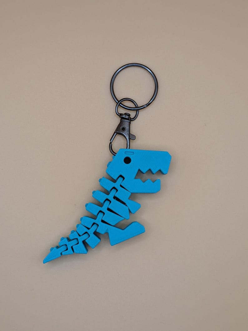 May include: A blue dinosaur skeleton keychain with a black metal clasp and keyring. The dinosaur skeleton is made of a segmented material, with a detailed rib cage, spine, and head. The keychain is a fun accessory.