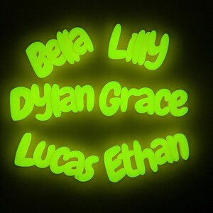 May include: A dark background highlights glowing green names: "Bella," "Lilly," "Dylan Grace," "Lucas," and "Ethan." The names are written in a bright, neon-like font, giving off a vibrant, luminous effect.